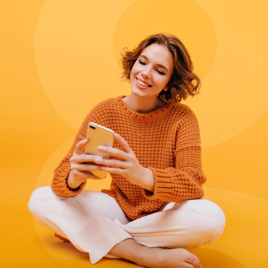 Mulher sentada no chão sorrindo enquanto usa o celular